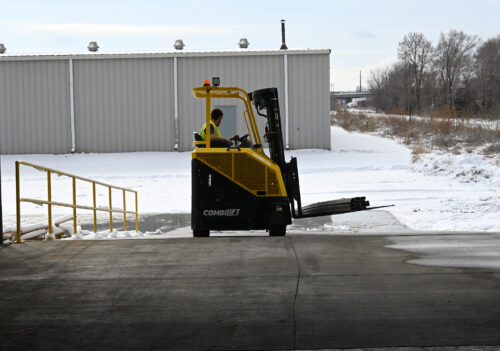 Operator driving a yellow CombiLift forklift outdoors on a snowy loading area near an industrial building.